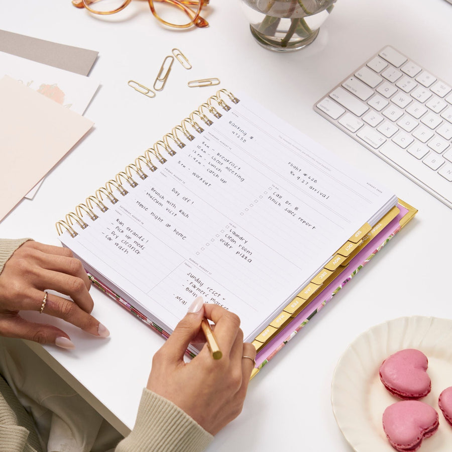 open book planner, written on page, gold binding, tabs, pen, and paper clips, dish with pink heart soaps, hands, white keyboard