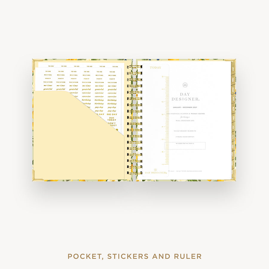 An open spiral-bound planner or agenda with a floral-patterned cover, showing a daily planner page on the right and a pocket section with stickers on the left. The text below reads 'POCKET, STICKERS AND RULER'.