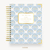 A spiral-bound planner or notebook shown from the front on a light neutral background. The cover has a repeating ornate fan-like pattern in light blue and gold on a white base, with a central gold label reading 'DAY DESIGNER.