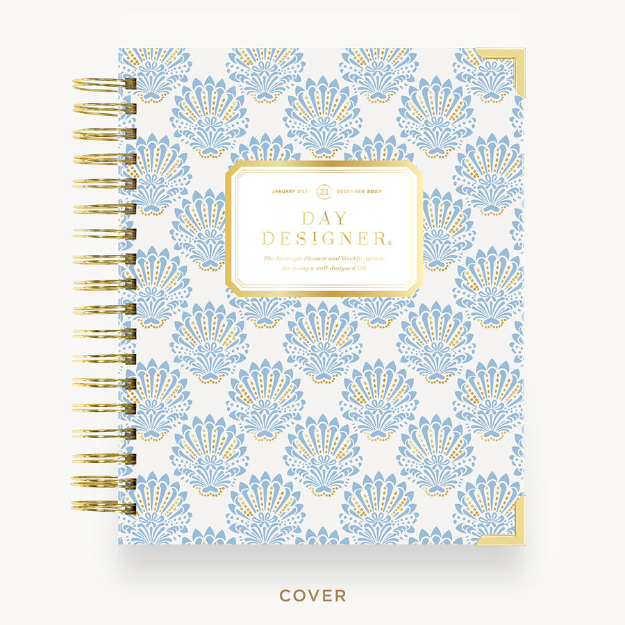 A spiral-bound planner or notebook shown from the front on a light neutral background. The cover has a repeating ornate fan-like pattern in light blue and gold on a white base, with a central gold label reading 'DAY DESIGNER.