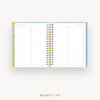 An open spiral-bound bucket list planner shown from above on a light gray background, with lined pages, gold rings, and a light blue and gold cover visible along the left and right edges. The text 'BUCKET LIST' appears below the planner.