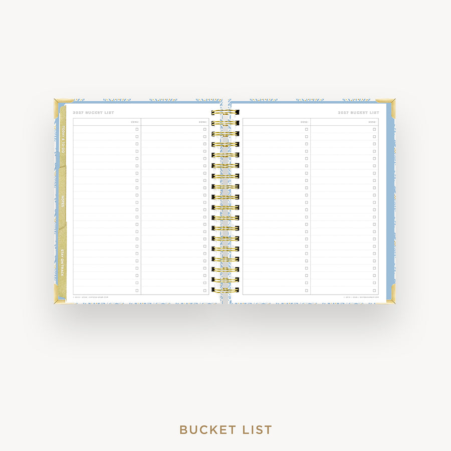 An open spiral-bound bucket list planner shown from above on a light gray background, with lined pages, gold rings, and a light blue and gold cover visible along the left and right edges. The text 'BUCKET LIST' appears below the planner.