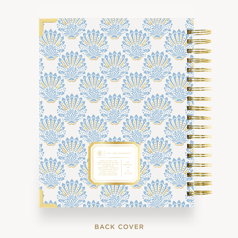 A spiral-bound planner or notebook shown from the back cover, featuring a repeating light blue and gold ornamental pattern on a white background, with a gold spiral binding and a small gold-bordered label area near the lower center.
