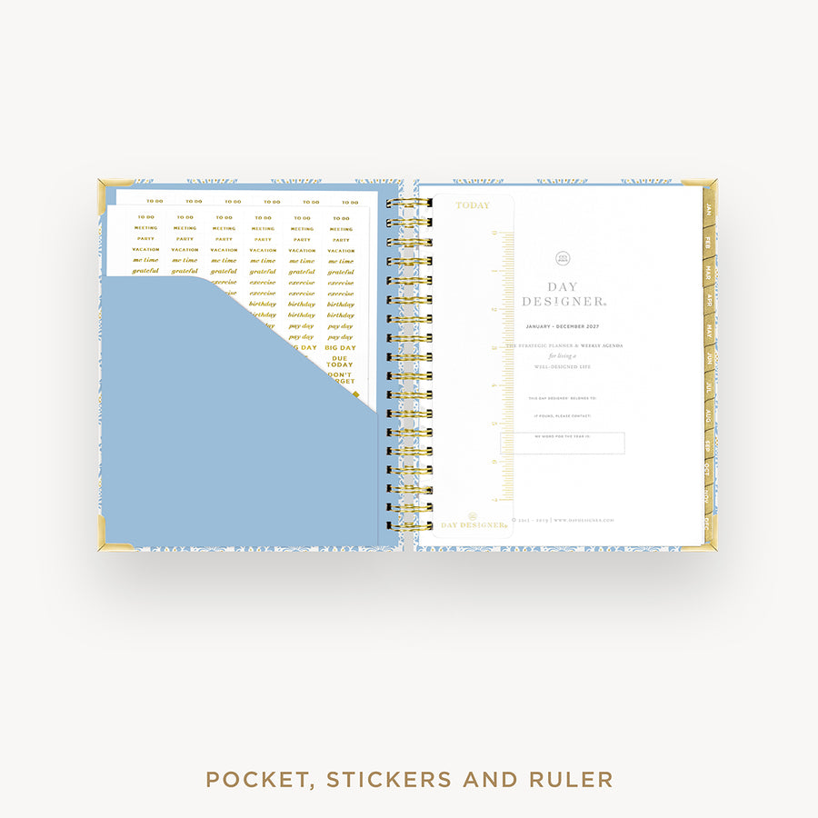 An open spiral-bound planner or organizer shown from above on a light gray background. The pages are white with gold text and layout elements, and a large light blue pocket or insert is visible on the left page.