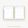 An open spiral-bound planner or notebook showing goal-setting worksheets on the pages, with a gold-toned ring binder and decorative gold and blue accents on the cover, photographed on a light gray background. Text below reads 'GOAL-SETTING WORKSHEETS'.