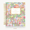 A spiral-bound Day Designer planner shown from the front cover, featuring a colorful watercolor illustration of European-style buildings and flowers. The cover has a cream label in the center with gold text, and the spiral binding is gold on the left.