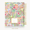 A spiral-bound planner or notebook shown from the back, featuring a colorful watercolor illustration of a European town with flowers, houses, and greenery. The right side shows a gold spiral binding, and a small label is visible near the bottom center.