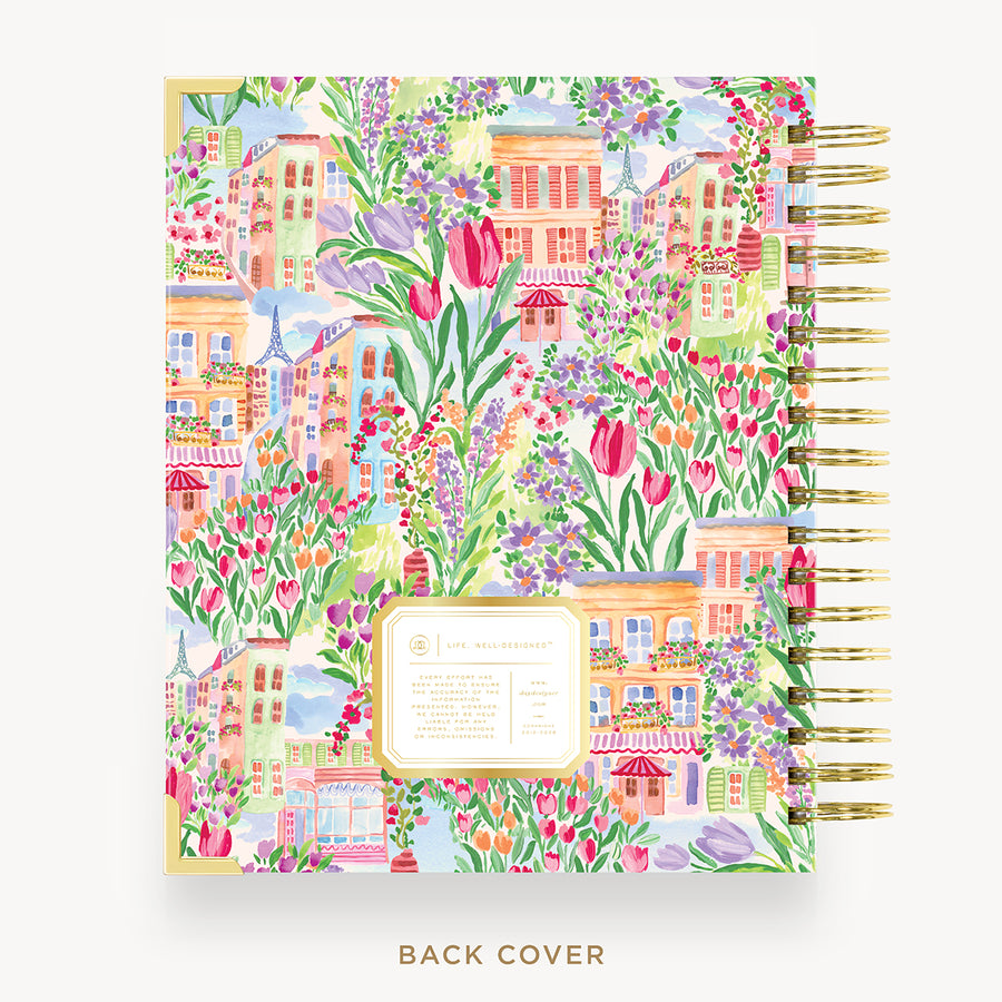 A spiral-bound planner or notebook shown from the back, featuring a colorful watercolor illustration of a European town with flowers, houses, and greenery. The right side shows a gold spiral binding, and a small label is visible near the bottom center.