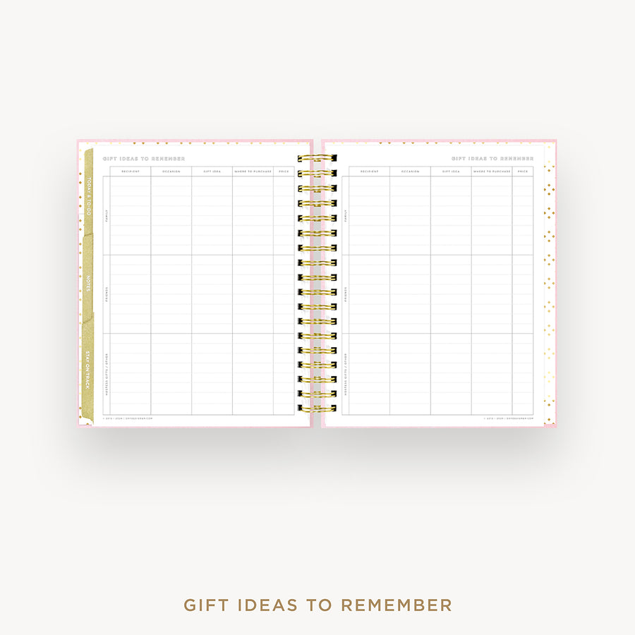 An open spiral-bound planner or notebook showing a gift ideas to remember tracking page, with a gold and black spiral binding, pink dotted borders, and a white page layout on a light gray background with the text 'GIFT IDEAS TO REMEMBER' below.
