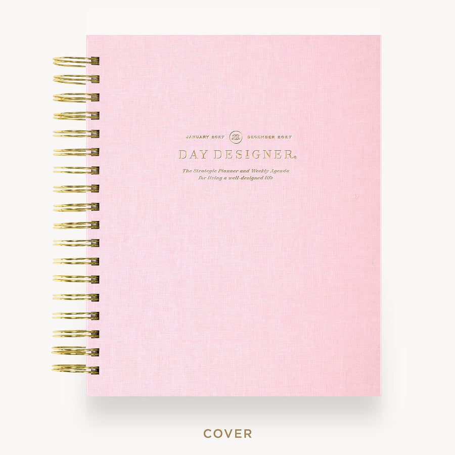 A light pink textured spiral notebook with gold wire binding and gold foil text on the cover reading 'DAY DESIGNER.' shown on a clean white background with the word 'COVER' below.