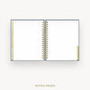 An open spiral-bound planner or notebook with lined pages, shown on a light gray background. The cover is dark navy blue, and the page edges and tab accents are a warm beige.