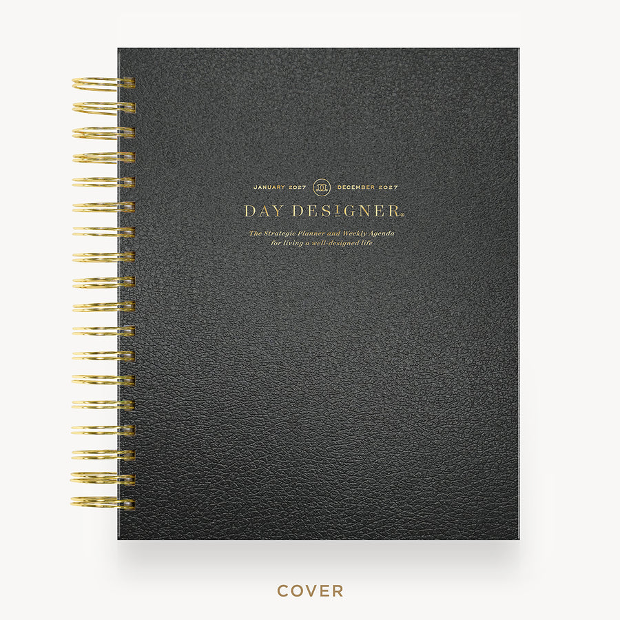 A black textured spiral-bound planner or agenda shown from the front on a light gray background. The cover has gold text reading 'DAY DESIGNER' and gold spiral binding along the left edge.