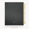 A black textured notebook or planner shown from the back cover, with a gold spiral binding along the right edge and a small gold circular logo near the bottom. The text 'BACK COVER' appears below on a light gray background.
