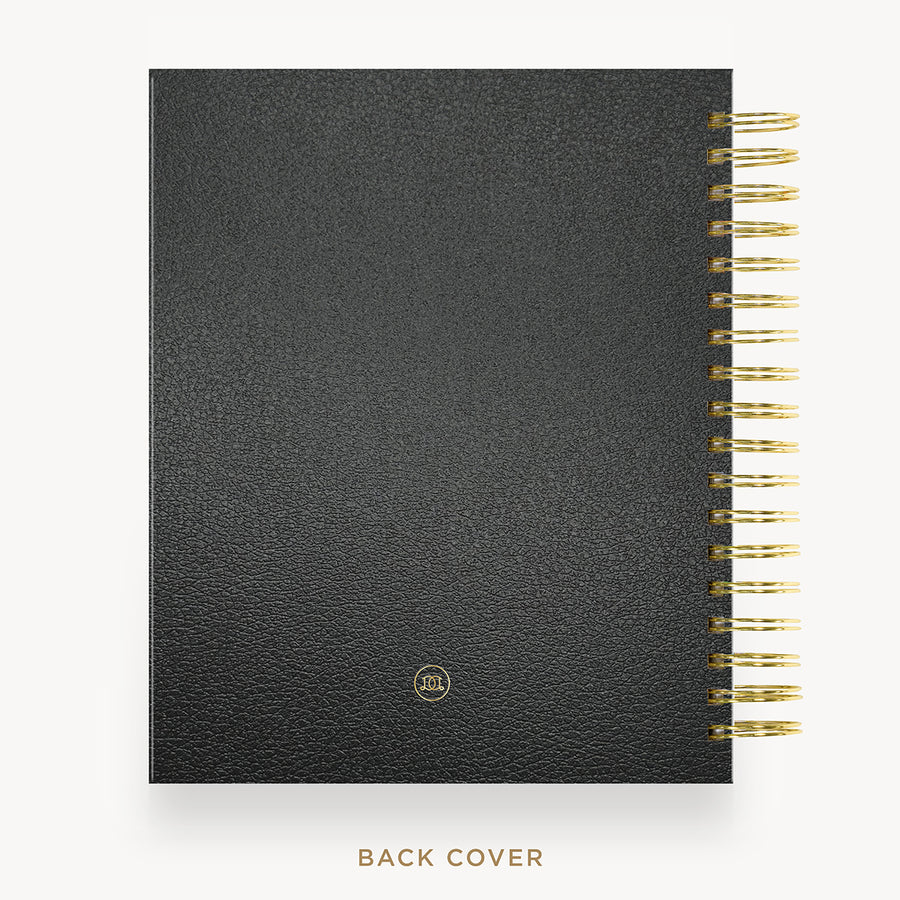 A black textured notebook or planner shown from the back cover, with a gold spiral binding along the right edge and a small gold circular logo near the bottom. The text 'BACK COVER' appears below on a light gray background.