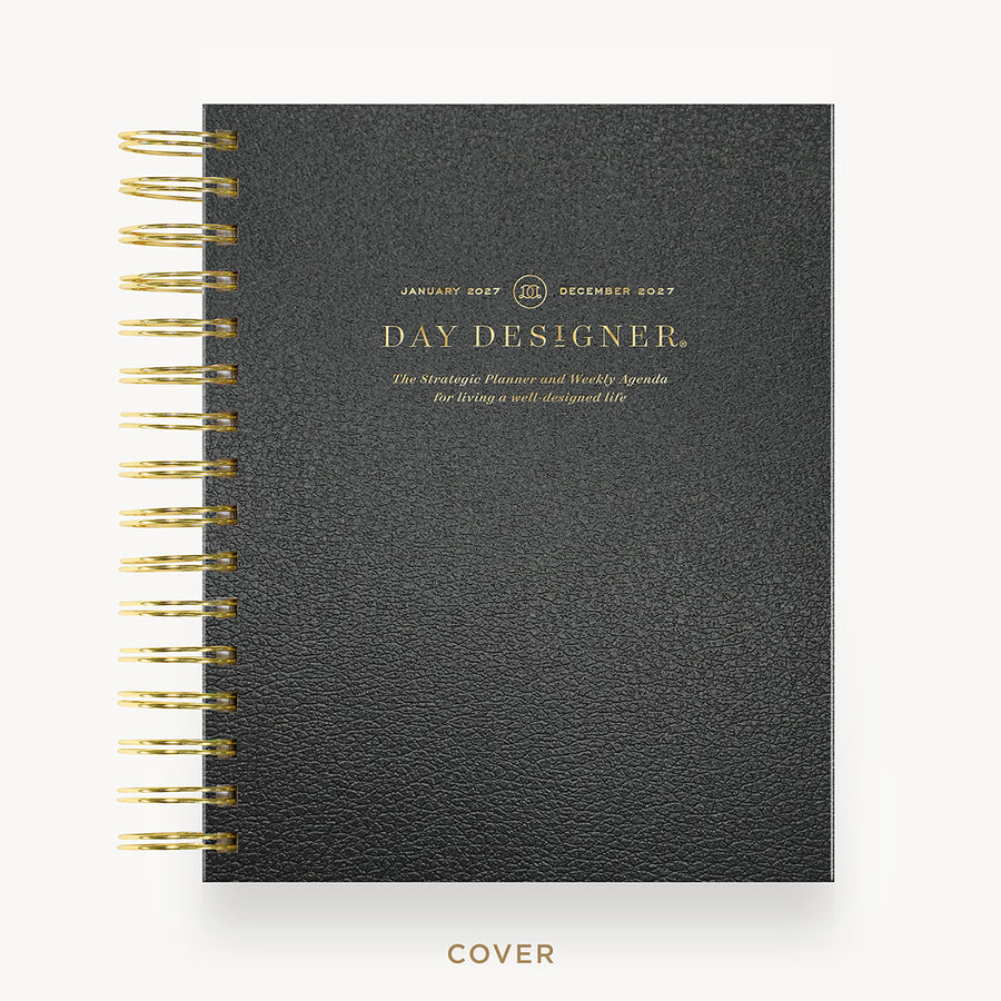 A black textured spiral-bound planner or journal shown closed, with gold wire binding and gold foil text on the cover reading 'DAY DESIGNER.' The cover is centered on a light gray background, with the word 'COVER' printed below.