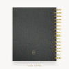 A black textured leather planner or notebook shown from the back, with gold spiral binding along the right edge and a small gold circular logo near the bottom. The text 'BACK COVER' appears below on a light gray background.