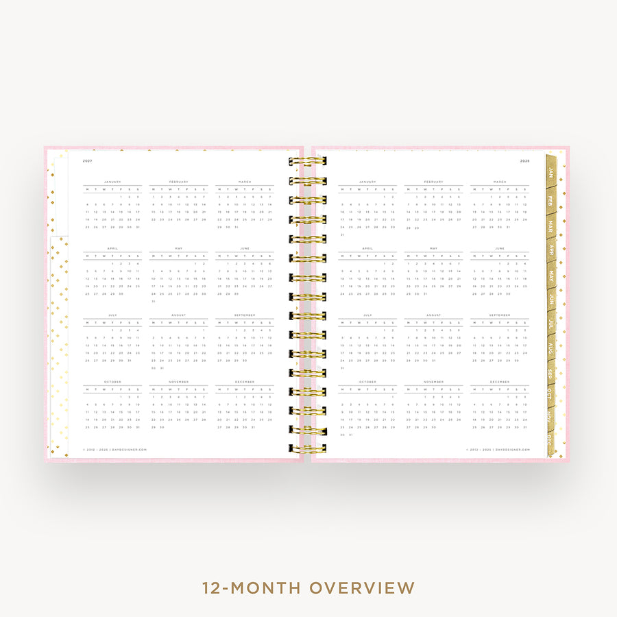 An open spiral-bound planner showing a 12-month calendar overview layout on white pages with a light pink border and gold accents, centered on a light gray background with the text '12-MONTH OVERVIEW' below.