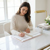 Woman writing in an open tabbed monthly calendar planner shown from the front, with January 2027 visible on the right page and a gold spiral binding down the center. The pages are white with light gray grid headers and subtle gold confetti accents on the left margin, desk and office background