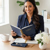 A woman in a navy blazer sits at a wooden desk holding an open navy planner with gold detailing. The desk is styled with a white mug, a small navy jewelry box, gold pens, and a vase of white flowers in a bright, modern office setting.
