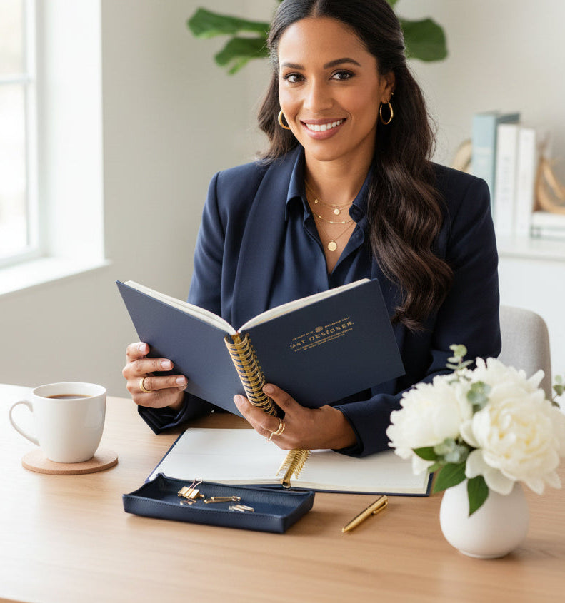 A woman in a navy blazer sits at a wooden desk holding an open navy planner with gold detailing. The desk is styled with a white mug, a small navy jewelry box, gold pens, and a vase of white flowers in a bright, modern office setting.