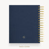 A navy blue fabric-covered notebook or planner shown from the back, with gold spiral binding along the right edge and a small circular logo near the lower center. The text 'BACK COVER' appears below on a light gray background.
