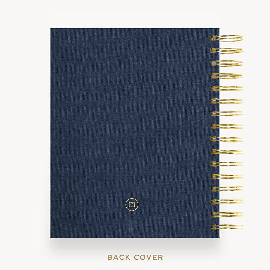 A navy blue fabric-covered notebook or planner shown from the back, with gold spiral binding along the right edge and a small circular logo near the lower center. The text 'BACK COVER' appears below on a light gray background.