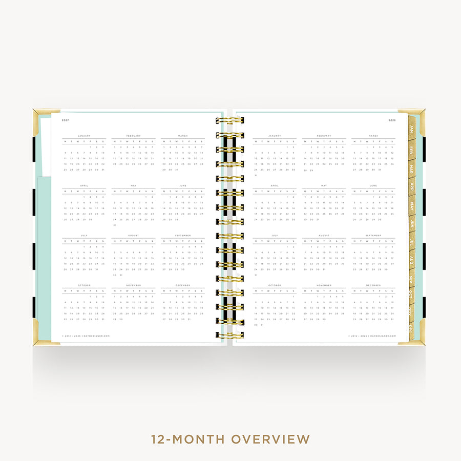 An open 12-month overview planner spread on a light gray background. The pages are white with black grid layouts for each month, held together by a central gold and black spiral binding.