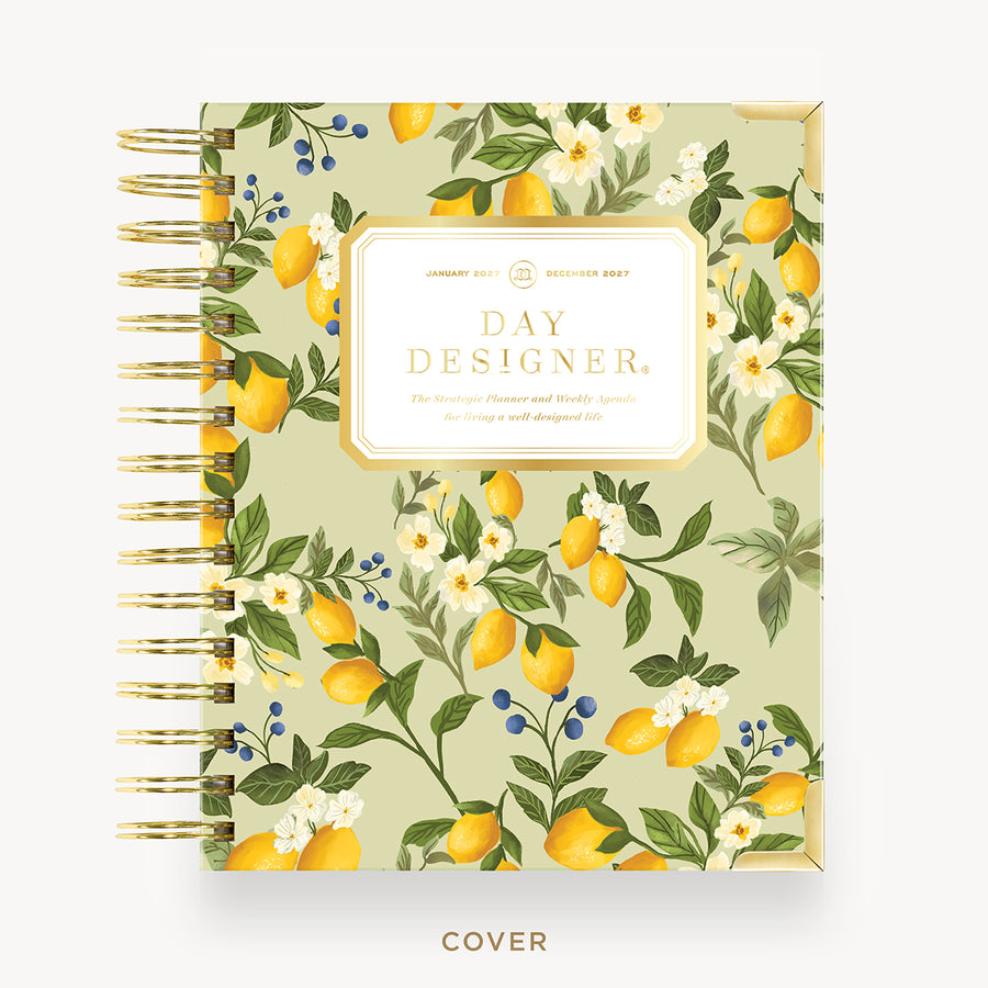 A Day Designer spiral-bound planner shown closed on a light beige background. The cover features a botanical pattern with yellow lemons, white flowers, green leaves, and small blue berries on a pale sage green base.