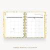 An open spiral-bound monthly calendar planner on a light gray background. The pages are white with a floral-patterned gold and green cover, and the left page shows tabs.