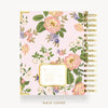 A floral-bound notebook shown from the back cover, featuring a light pink background with large pink and white flowers, green leaves, and purple blossoms. The notebook has gold-toned spiral binding and a decorative gold-bordered label on the cover.