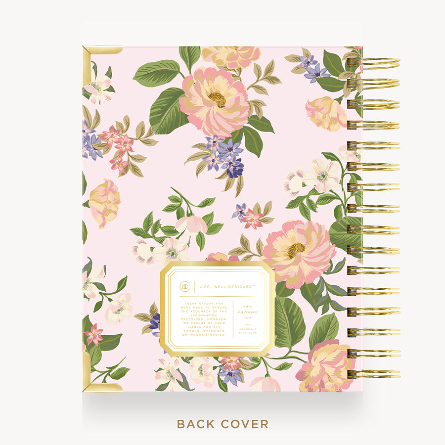 A floral-bound notebook shown from the back cover, featuring a light pink background with large pink and white flowers, green leaves, and purple blossoms. The notebook has gold-toned spiral binding and a decorative gold-bordered label on the cover.