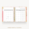 An open monthly calendar planner with two visible pages showing January 2027, bound by gold-tone wire loops. The cover has a pink base with floral accents, and the text 'TABBED MONTHLY CALENDAR' appears below on a light gray background.