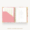 An open spiral planner shown from above on a light gray background. The planner has a floral-patterned cover in pink, white, and gold, with a pink pocket on the left, white sticky notes, and a white page with gold text on the right.