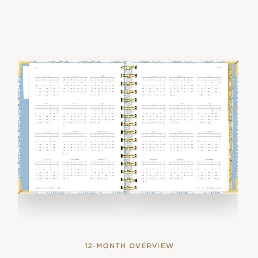 An open spiral-bound planner showing a 12-month overview calendar layout on white pages, with a light blue and pale yellow patterned cover and gold wire binding, centered on a light gray background with the text '12-MONTH OVERVIEW' below.