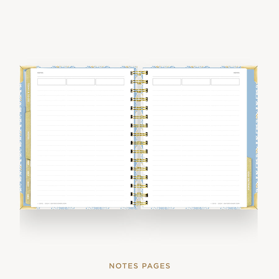An open spiral-bound planner or notebook with lined pages, shown on a light gray background. The cover and edges have a pale blue and white patterned design with gold-toned spiral binding.