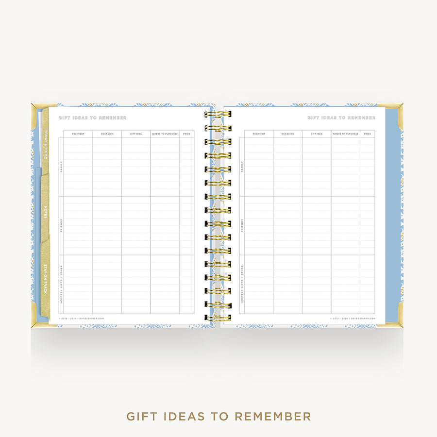 An open spiral-bound gift tracker planner on a light gray background. The pages are white with light blue borders and gold accents, featuring tables for recording gift ideas.