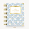 A Day Designer spiral-bound planner cover shown from the front on a light neutral background. The cover features a repeating light blue and gold fan-like pattern on a white base, with a gold-bordered central label reading 'DAY DESIGNER' and gold spiral binding along the left side.