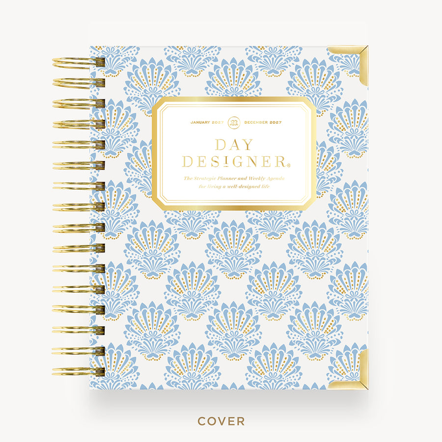 A Day Designer spiral-bound planner cover shown from the front on a light neutral background. The cover features a repeating light blue and gold fan-like pattern on a white base, with a gold-bordered central label reading 'DAY DESIGNER' and gold spiral binding along the left side.