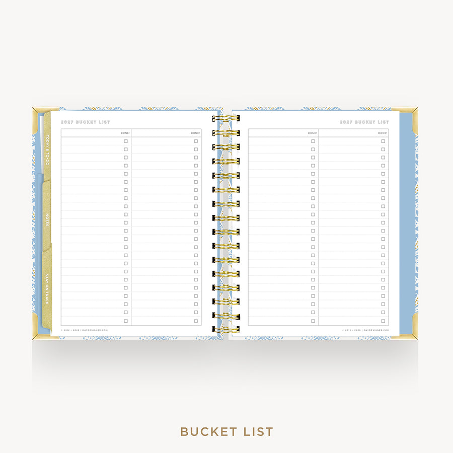 An open spiral-bound bucket list planner on a light gray background. The pages are lined with check boxes and labeled 'BUCKET LIST' items, with a gold spiral binding and a light blue and white patterned cover.