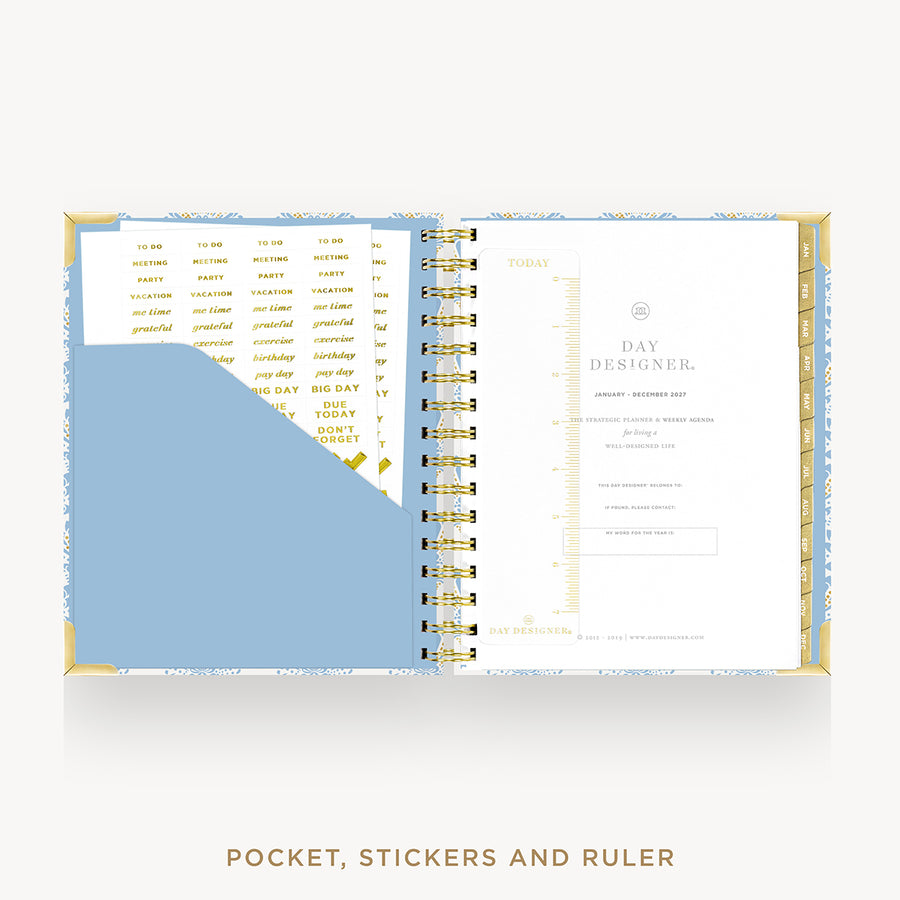 An open spiral-bound planner or pocket organizer shown from above on a light gray background. The product includes a light blue pocket, gold-and-white printed inserts, a white page with gold text, and a ruler.