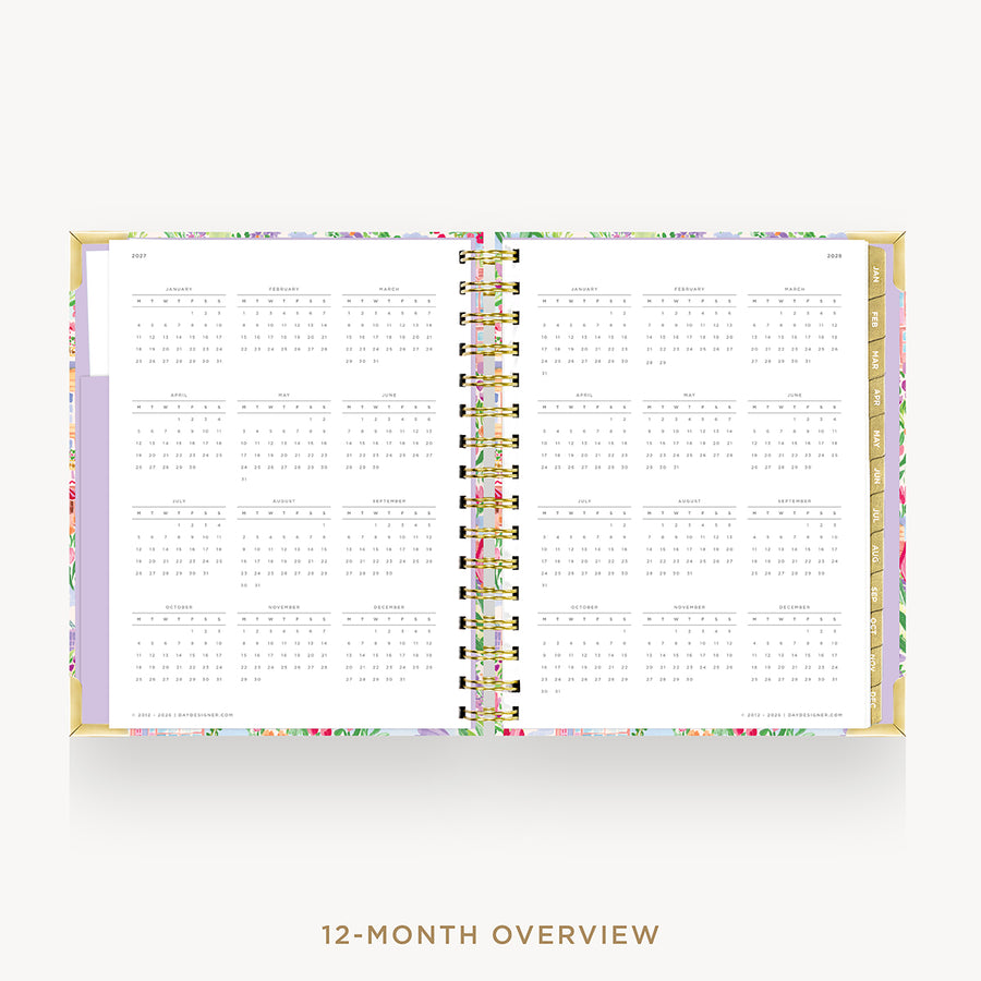 An open 12-month overview planner with a white interior page layout, a pastel floral patterned cover, and gold-toned spiral binding, shown on a light gray background with the text '12-MONTH OVERVIEW' below.