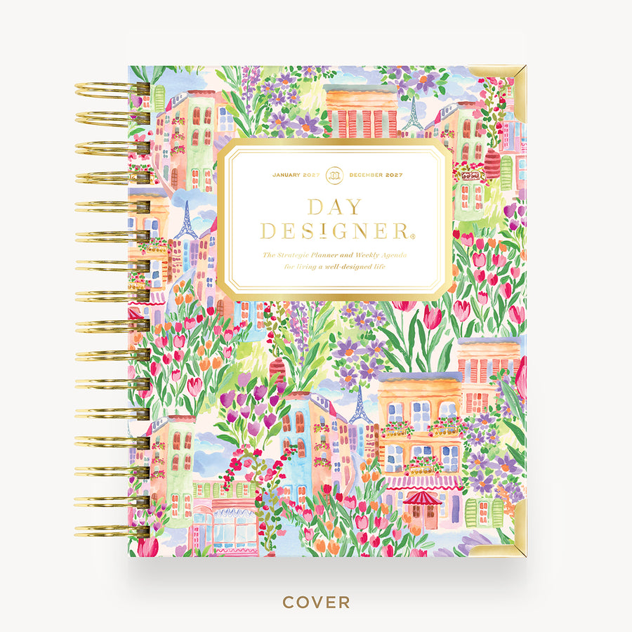 A Day Designer January 2027 planner, featuring a colorful watercolor illustration of Parisian houses, flowers, and greenery, with gold spiral binding and a cream title label.
