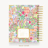 A spiral-bound planner or notebook shown from the back, featuring a colorful watercolor illustration of a European town with flowers, tulips, and pastel buildings. The cover has a gold border and a central cream label area.