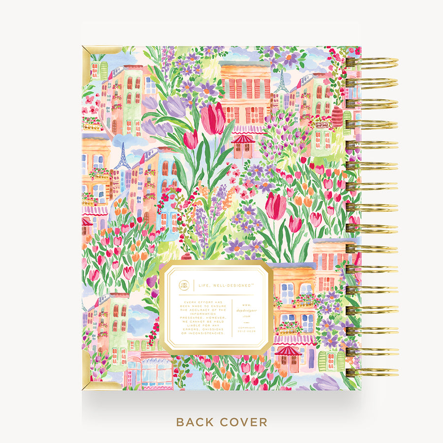 A spiral-bound planner or notebook shown from the back, featuring a colorful watercolor illustration of a European town with flowers, tulips, and pastel buildings. The cover has a gold border and a central cream label area.