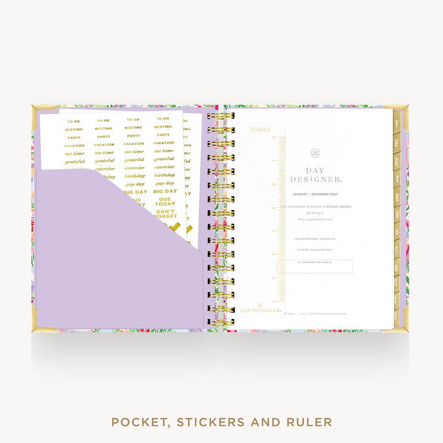 An open spiral-bound planner shown from above on a white background, with a floral-patterned cover, gold-toned ring binder, lavender pocket, and printed inserts. Text below reads 'POCKET, STICKERS AND RULER'.