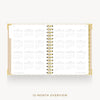 An open spiral-bound planner or calendar showing a 12-month overview layout on white pages, with a gold-toned binding and a light beige cover edge, photographed on a clean light background.