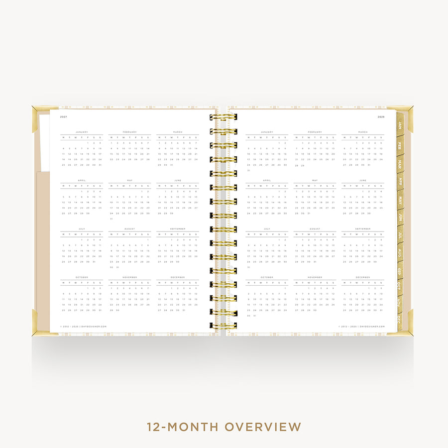 An open spiral-bound planner or calendar showing a 12-month overview layout on white pages, with a gold-toned binding and a light beige cover edge, photographed on a clean light background.