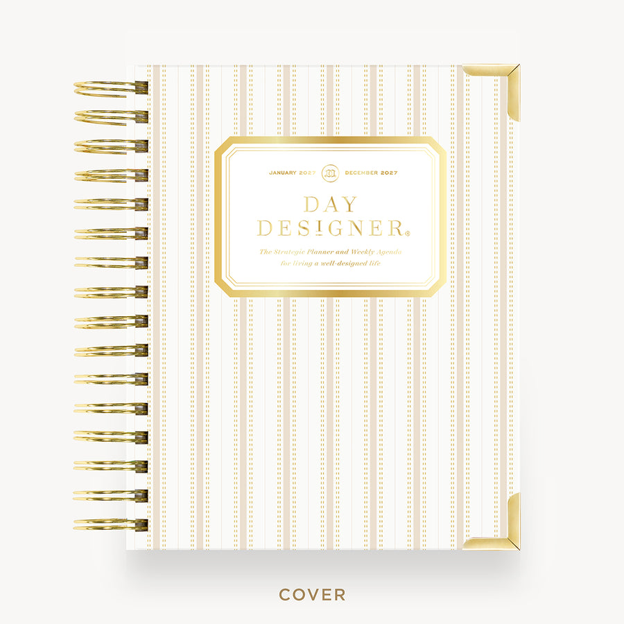 A Day Designer spiral notebook with a cream and gold striped cover, featuring a gold-bordered label in the center and gold wire binding on the left, shown on a light gray background.