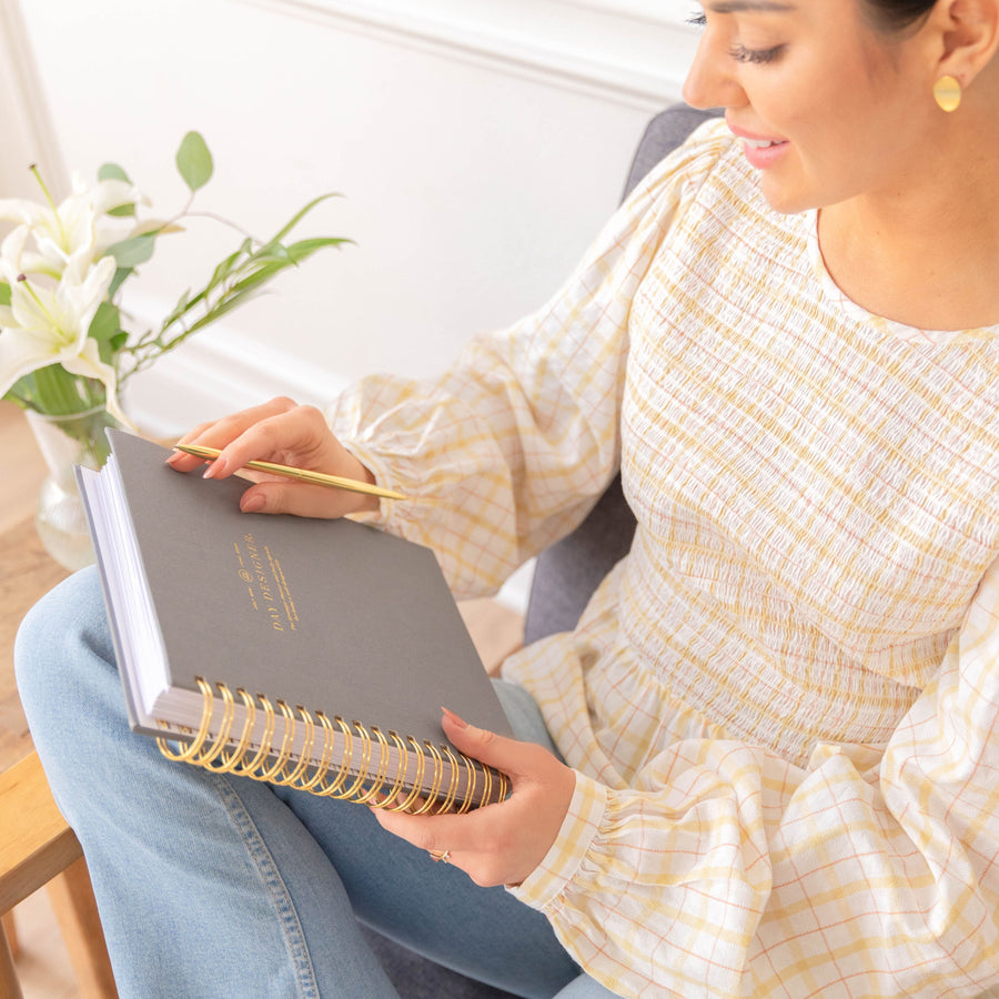 A woman in a cream and yellow checkered blouse and blue jeans holds a gray spiral notebook with gold lettering and a gold spiral binding, using a gold pencil. A vase of white lilies is visible in the background.