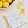 An open spiral-bound planner or notebook with gold rings lies on a light surface, showing handwritten notes on white pages. A glass of lemonade with lemon slices, a bowl of lemons, and a loose lemon are arranged around it as styled props.